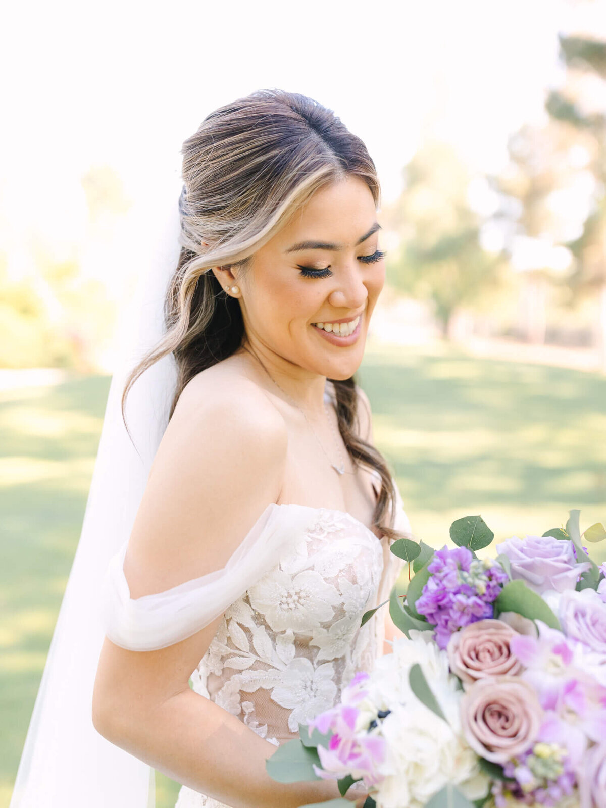 A bride in a white lace off-shoulder wedding dress smiles while holding a bouquet of purple and white flowers. Sunlit greenery forms the serene background.