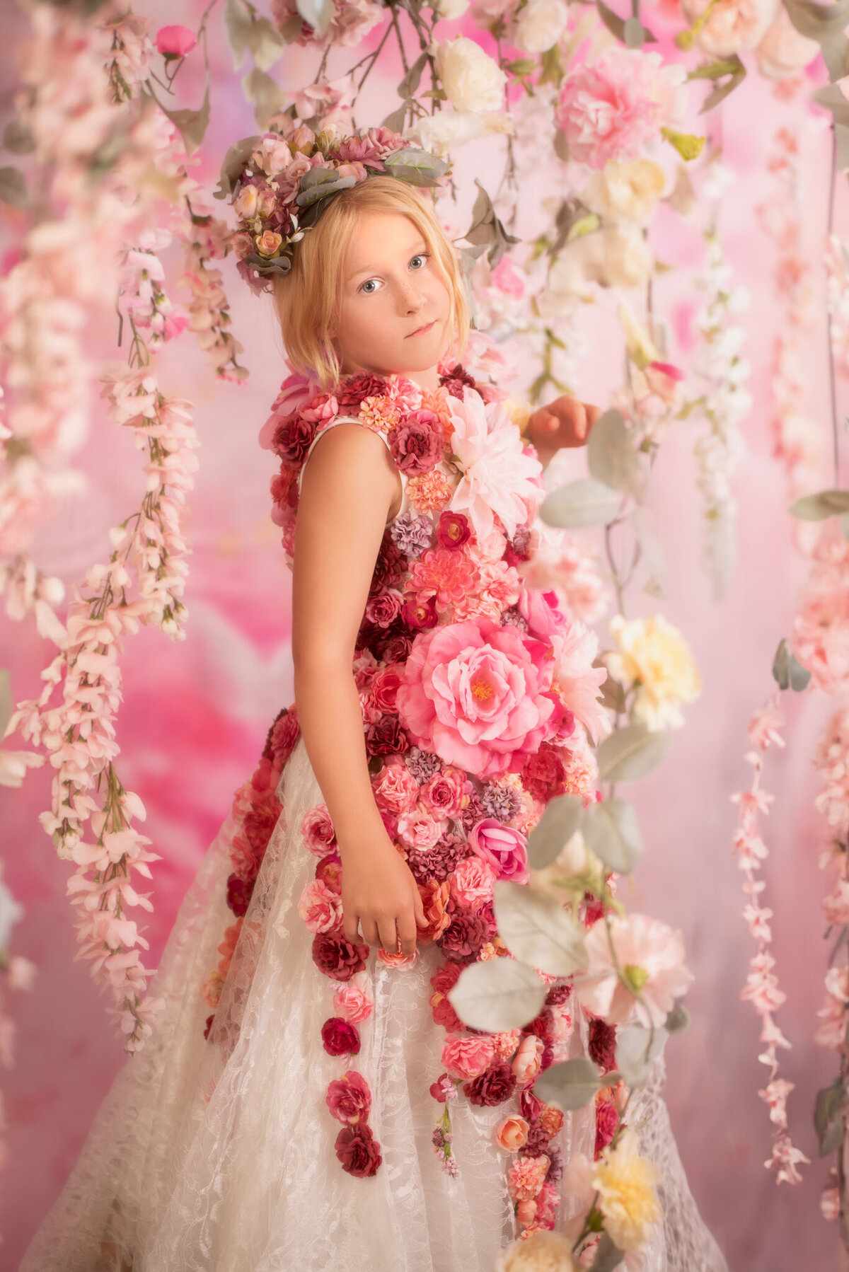 Whimsical portrait of a little girl on a swing adorned with pink flowers, wearing an elegant floral gown. Captured in Overland Park, Kansas, this enchanting setup is perfect for creative and artistic child photography.