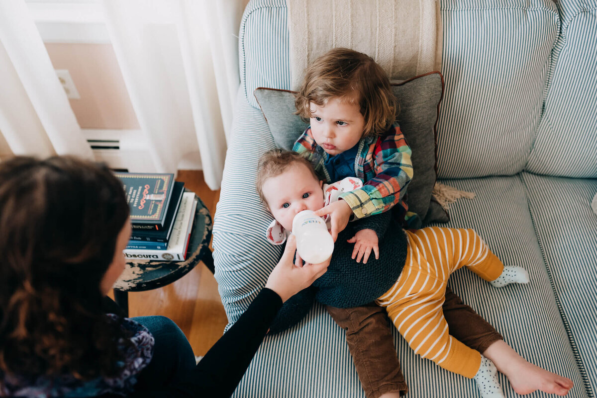 A big brother holds his baby sister in his lap and feeds her a bottle as his mother helps him