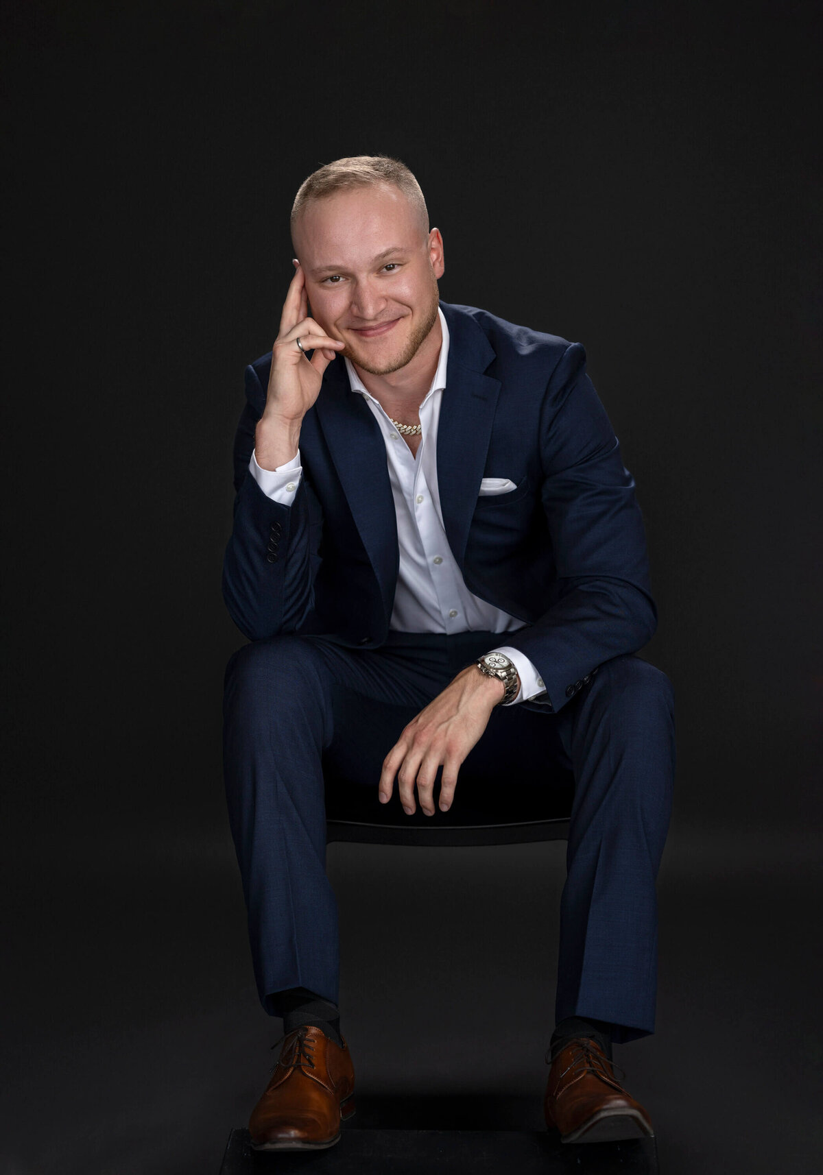 A young businessman in a navy suit, sitting with a relaxed yet professional posture against a dark backdrop.