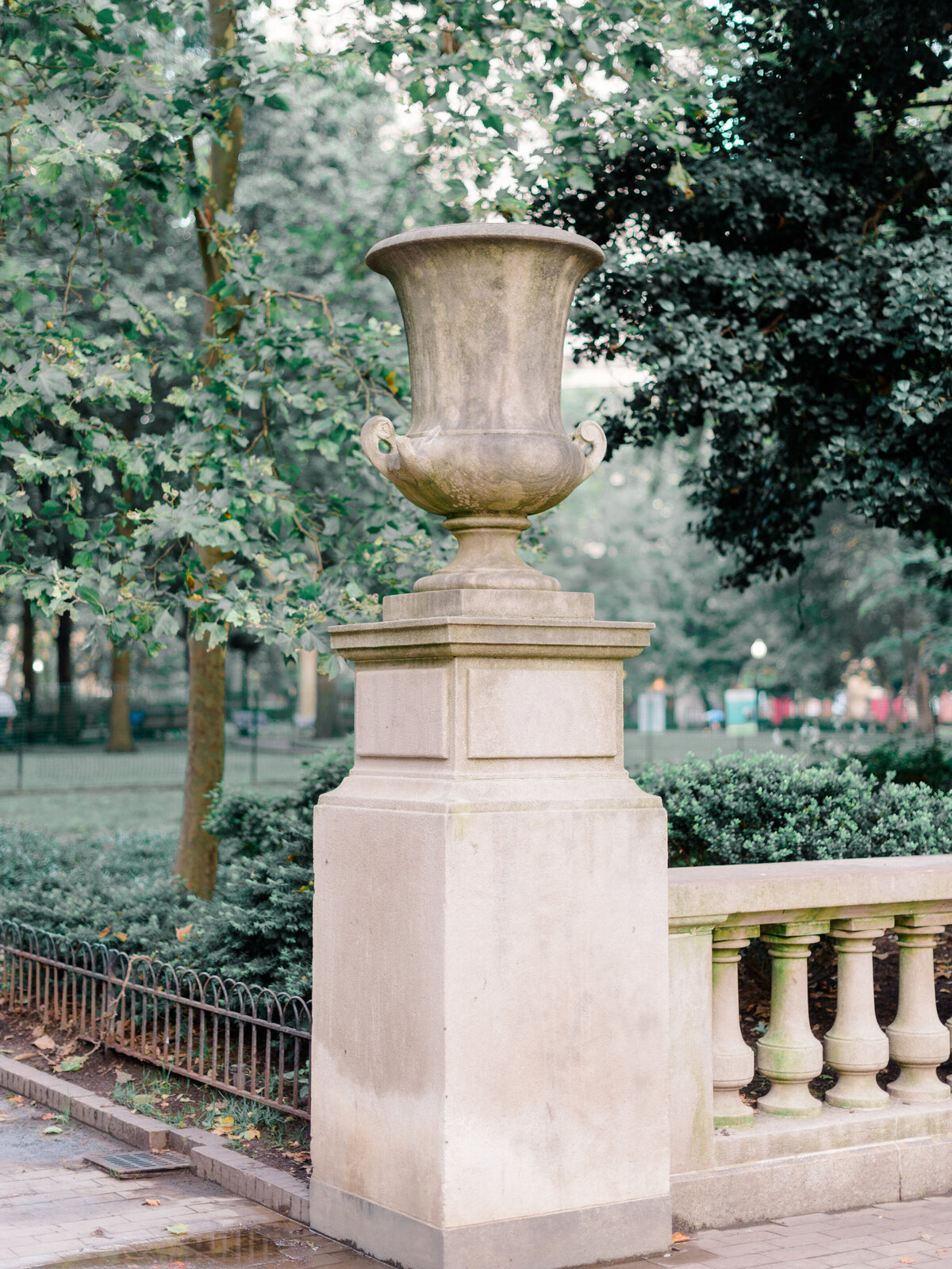 A stately stone urn atop a pedestal stands at the edge of Rittenhouse Square in Philadelphia, framed by lush greenery and historic balustrades. Captured in soft, natural light by Elizabeth Kane Photography, a Luxury Philadelphia Wedding Photographer.