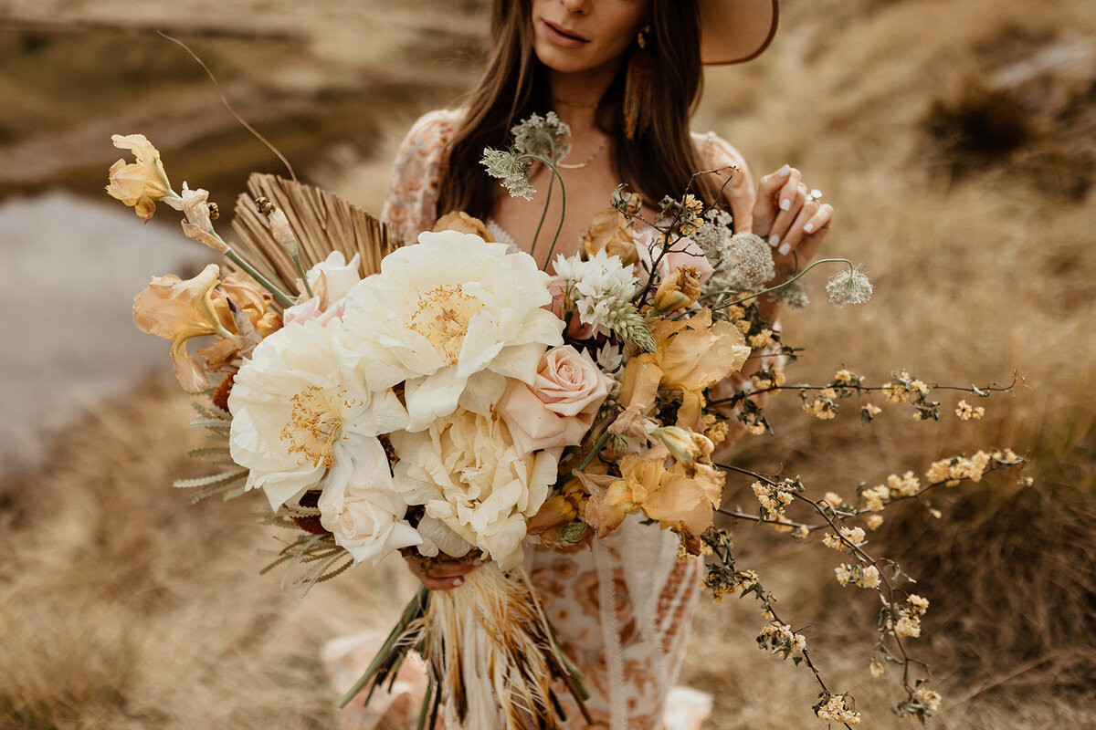 The Vase Floral Co - bride in Queenstown walks in field with large flower bouquet in pale yellows and whites