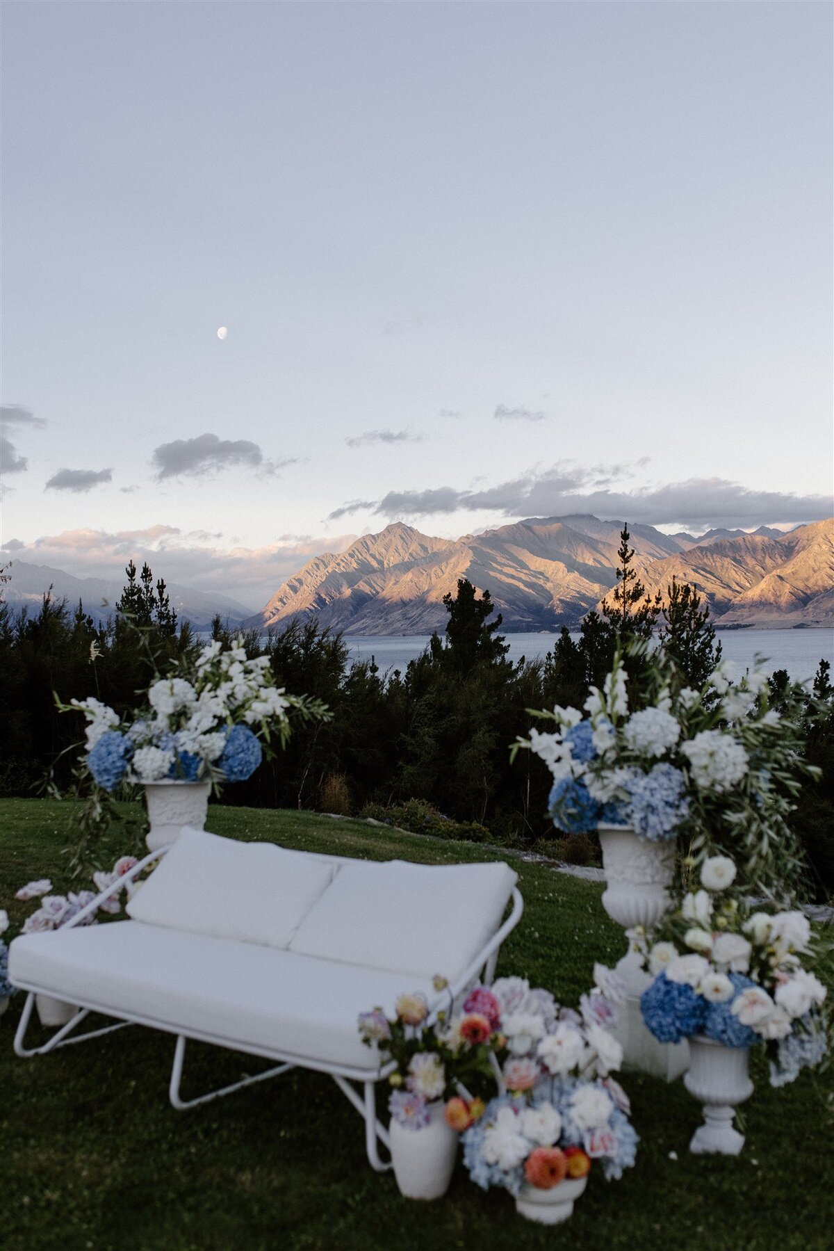White outdoor lounge set framed by urns and buckets of fresh pastel flowers, with a mountain and lake backdrop under a soft, dusky sky.
