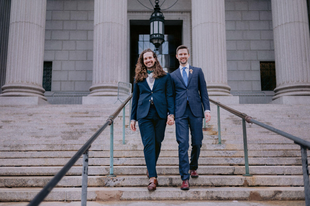 The two grooms are holding hands while standing on the grand staircase outside NYC City Hall. Elopement Image by Jenny Fu Studio