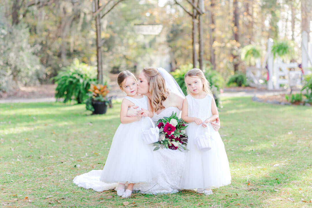 A bride leans down to kiss a flower girl on the cheek while taking photos.