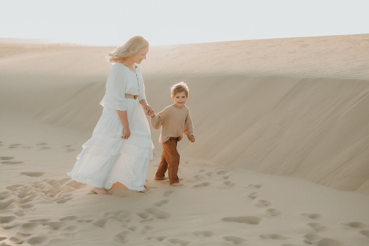 Woman in a flowing white dress walks with her young son and holds his hand as they walk along the beach at sunrise