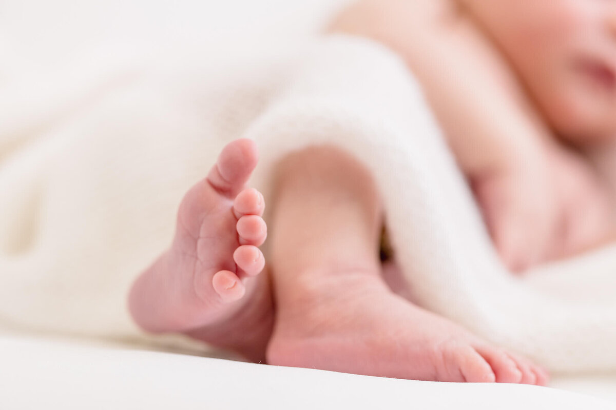 Close-up of a babys feet peeking out from under a soft white blanket. The scene is gently lit, emphasizing the tiny toes and softness of the blanket, creating a serene and peaceful atmosphere.