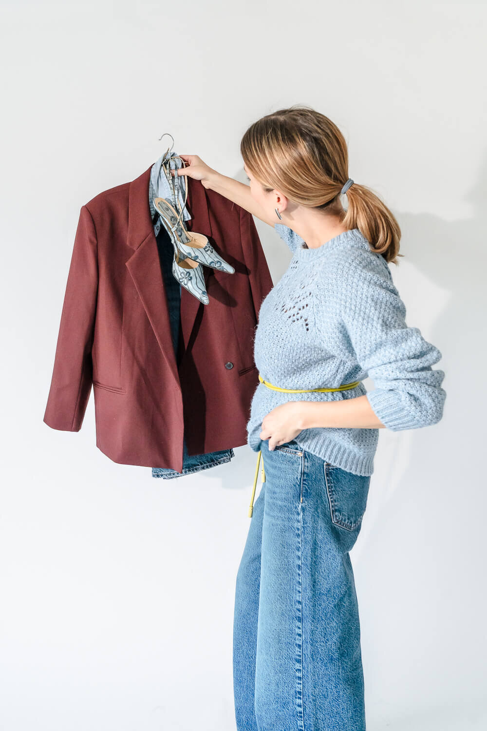 Woman in blue sweater and jeans choosing burgundy blazer and floral heels from clothing rack.