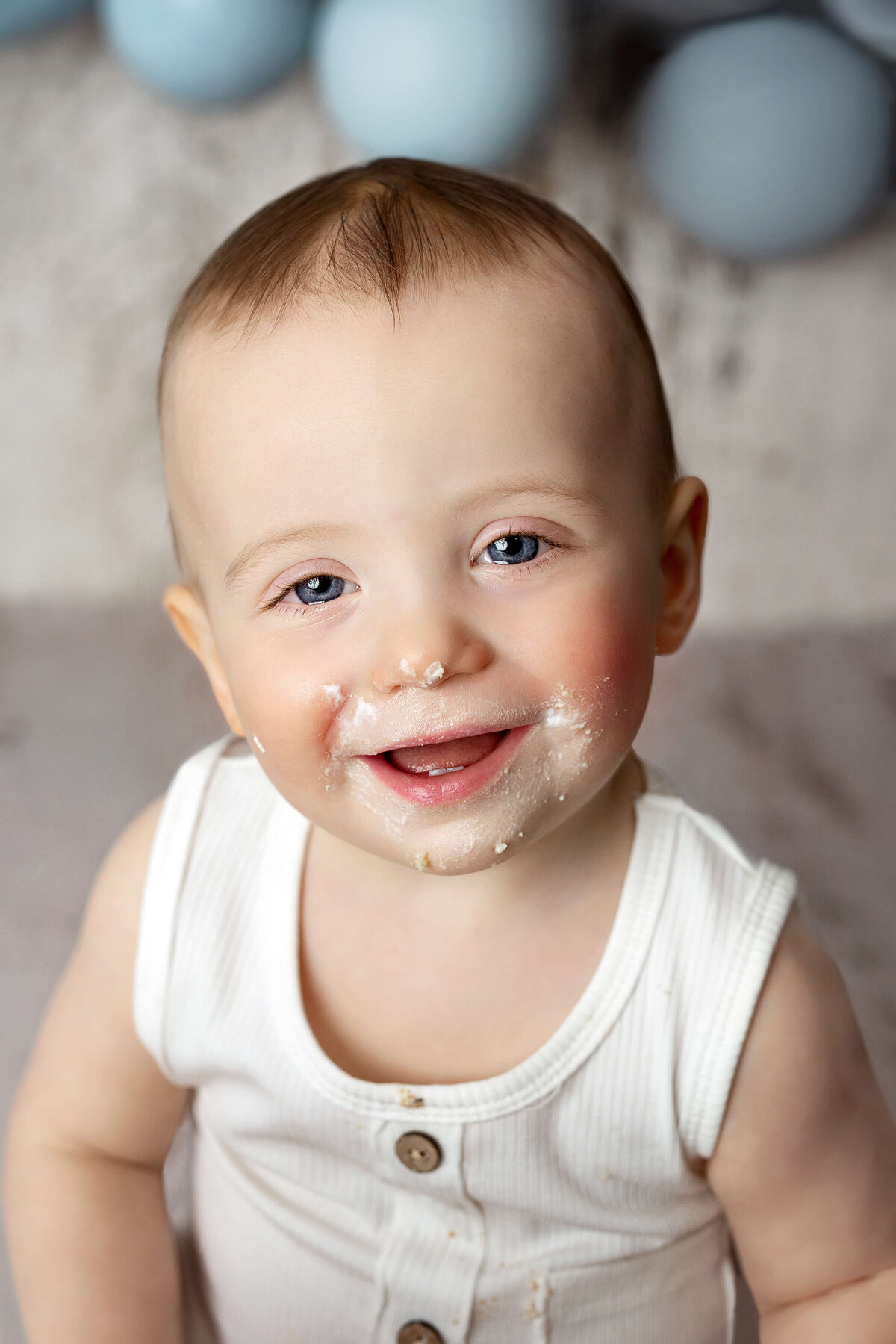 little boy with cake on his face smiling in a white outfit during his one year cake smash session