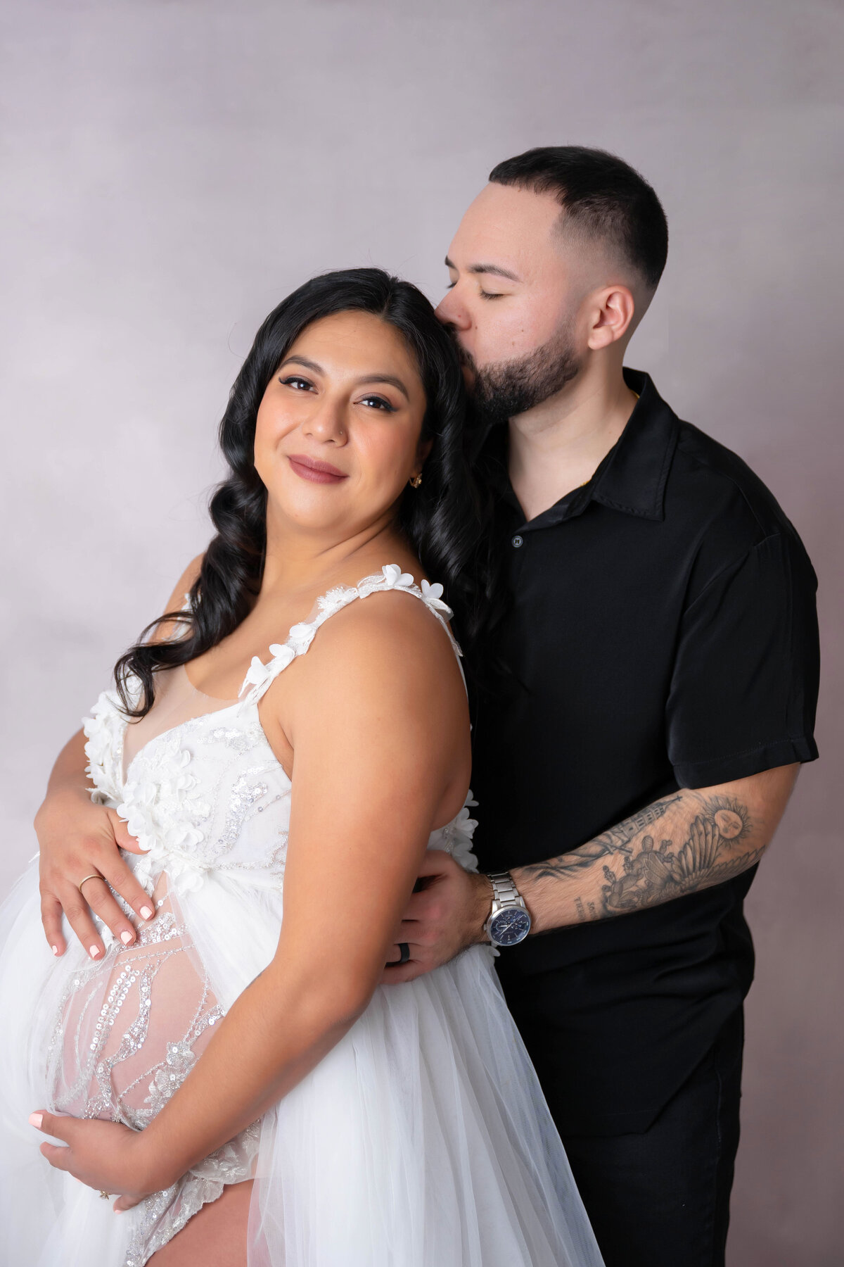 Pregnant woman holds belly while partner hugs her from behind and kisses her head.