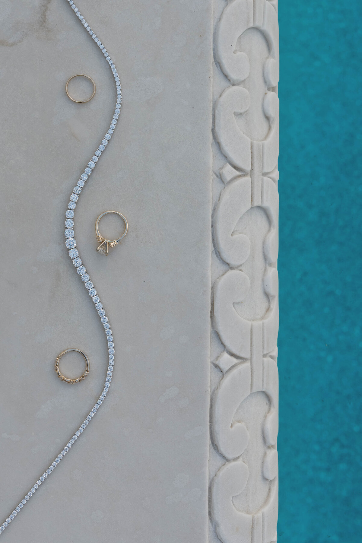 A fine art flat lay of wedding jewelry, including diamond rings and a delicate bracelet, set on a marble surface near a pool. A luxurious wedding detail shot with a refined aesthetic.