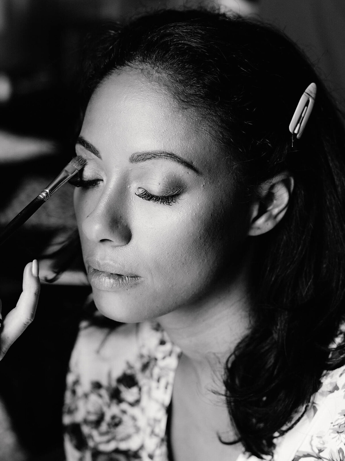 Black and white photo of a woman with eyes closed while an eye shadow is applied on her eyes