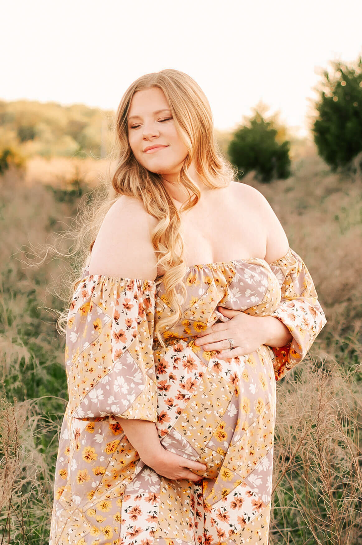 pregnant mom in floral dress hugging belly at a field during Branson MO maternity photography