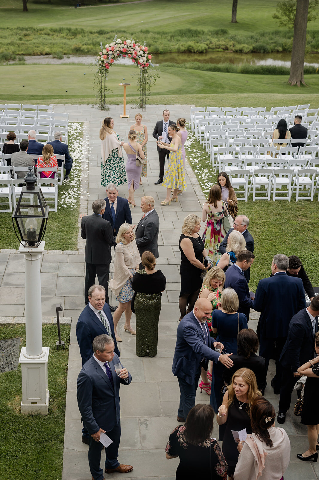 Guests in colorful spring attire mingling before sitting down in white chairs for wedding ceremony at Fiddler'S Elbow Country Club
