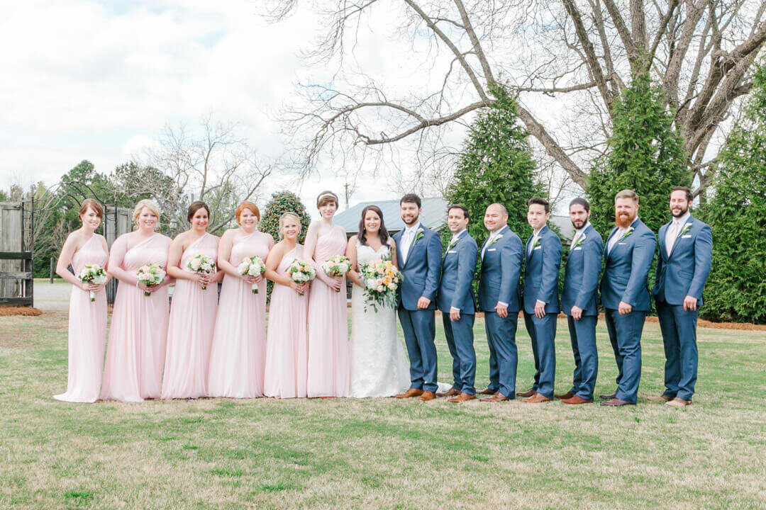 A bride and groom stand and smile with their large wedding party in a lawn