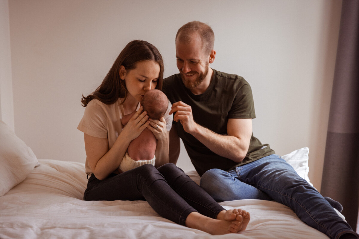 Livsstilsfoto familieportrett der foreldrene sitter på en seng; mor holder baby og kysser baby, far holder rundt mor og ser på baby.