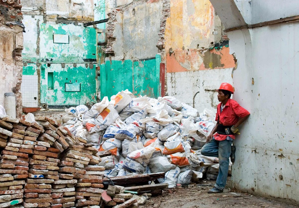 A construction worker leans on a wall to take a break with a large pile of construction materials in front of him. Social Documentary photo with narrative quality.