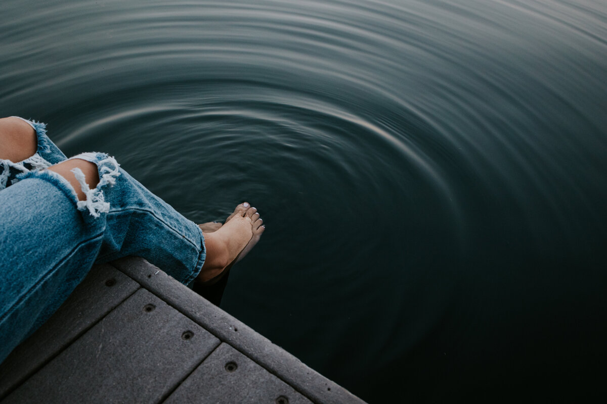 Young woman sits on dock while dipping toes in lake.