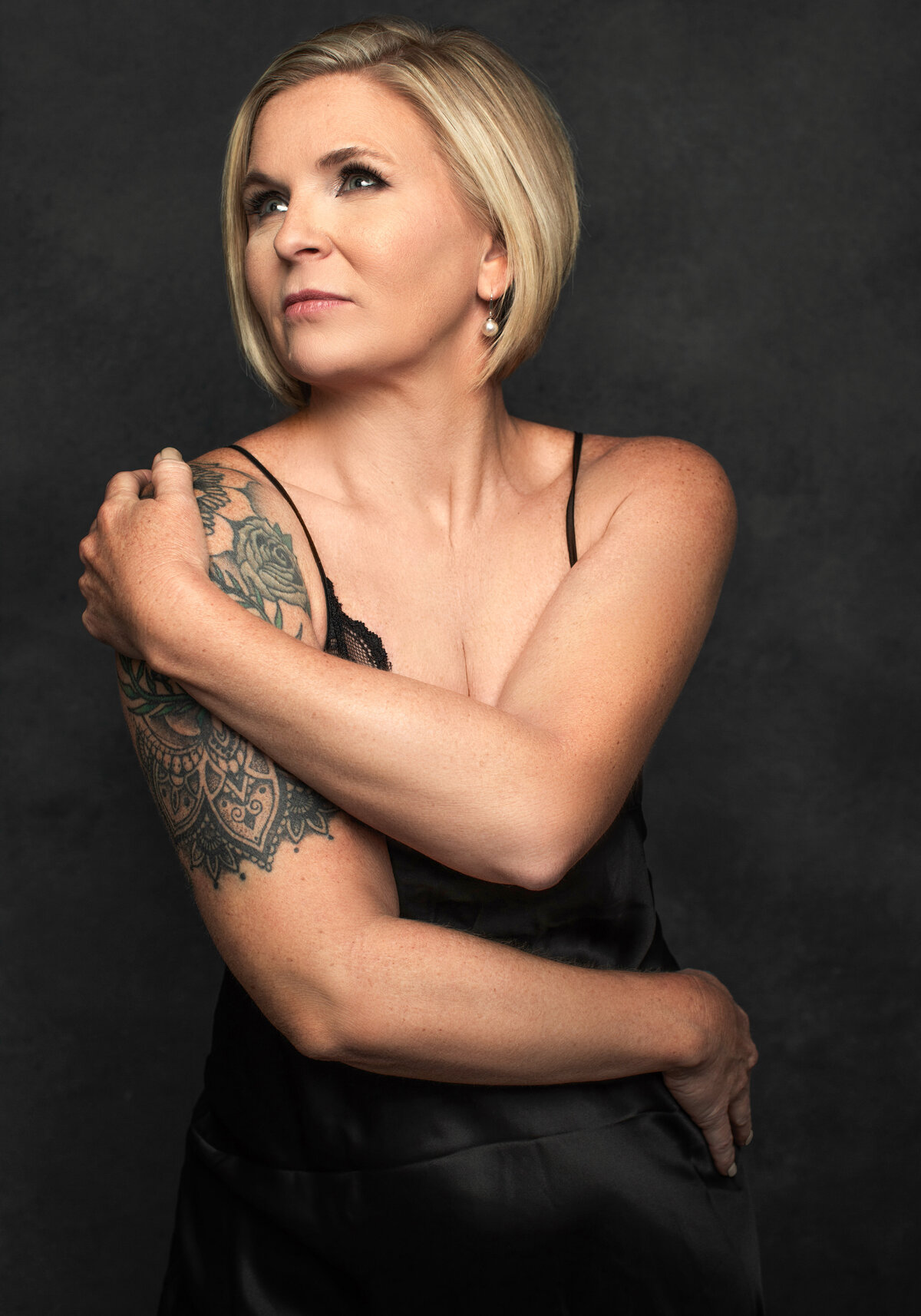 A woman with a blond pixie cut hairstyle embraces herself while wearing a black dress as she poses for a portrait photo at Janel Lee Photography studios in Cincinnati Ohio
