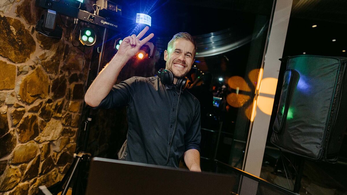 Male DJ smiling and flashing a peace sign behind the decks at a wedding reception with colourful lights.