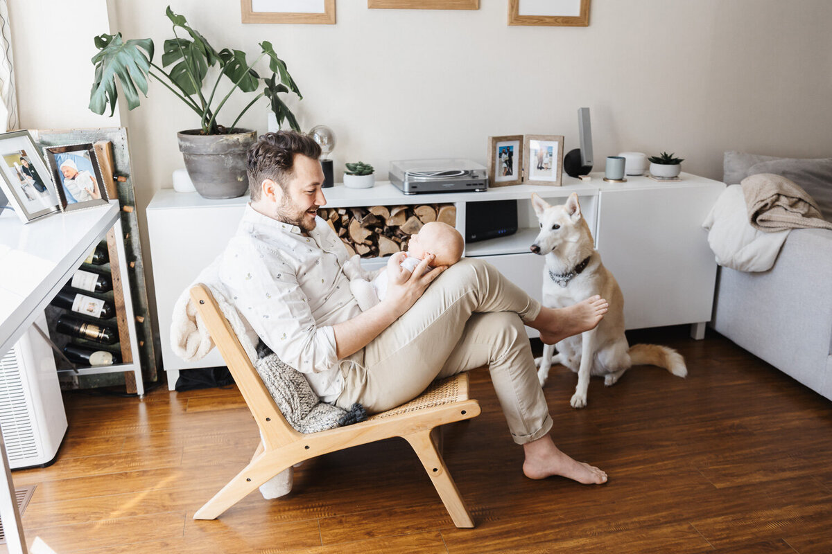 dad and newborn son sitting  on  chair in living room with pet dog looking on