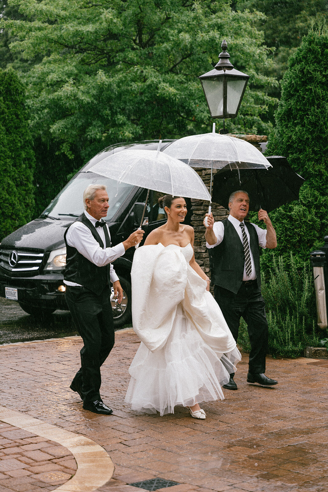 Bride is escorted to the Somerly at Fox Hollow by two gentlemen holding umbrellas in the rain.