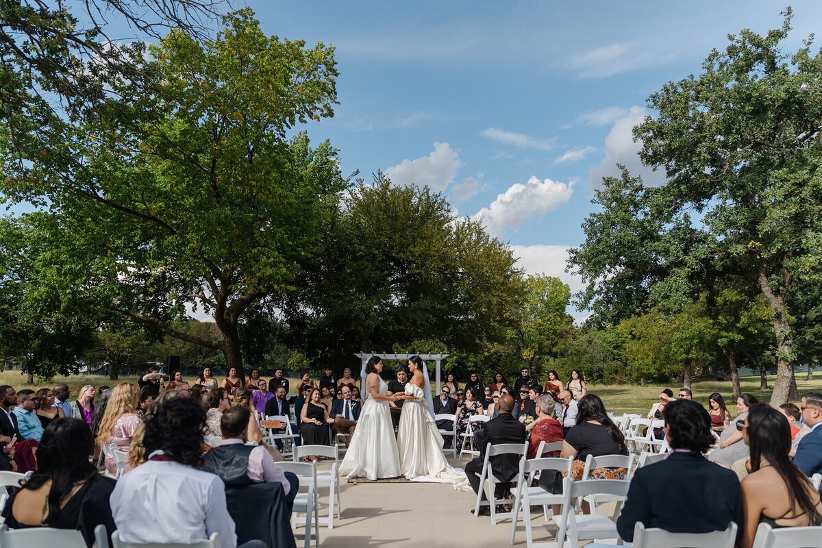 A candid shot of two brides during their wedding ceremony in Fort Worth, Texas.  Both brides are holding hands and facing towards each other; they are both wearing long, flowing, white dresses with long, white veils. Guests are seated in a large circle around them, and the whole scene is backed by many trees and a large blue sky.