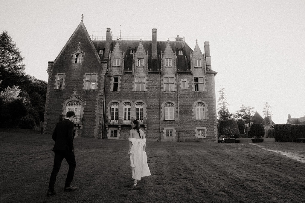 Mariés de dos marchant dans l'herbe en direction d'un grand château. Photographie en noir et blanc, par Léa, photographe mariage vintage.