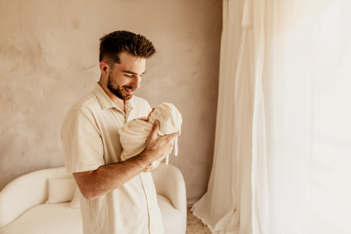 Father looks at his baby and smiles down at her near a window.