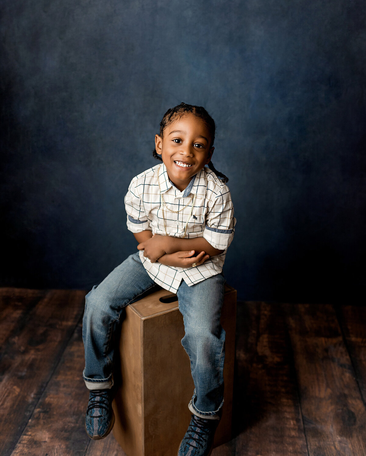 A boy with braided hair  sitting on a  box with arms folded and smiling during Brooklyn child portrait session