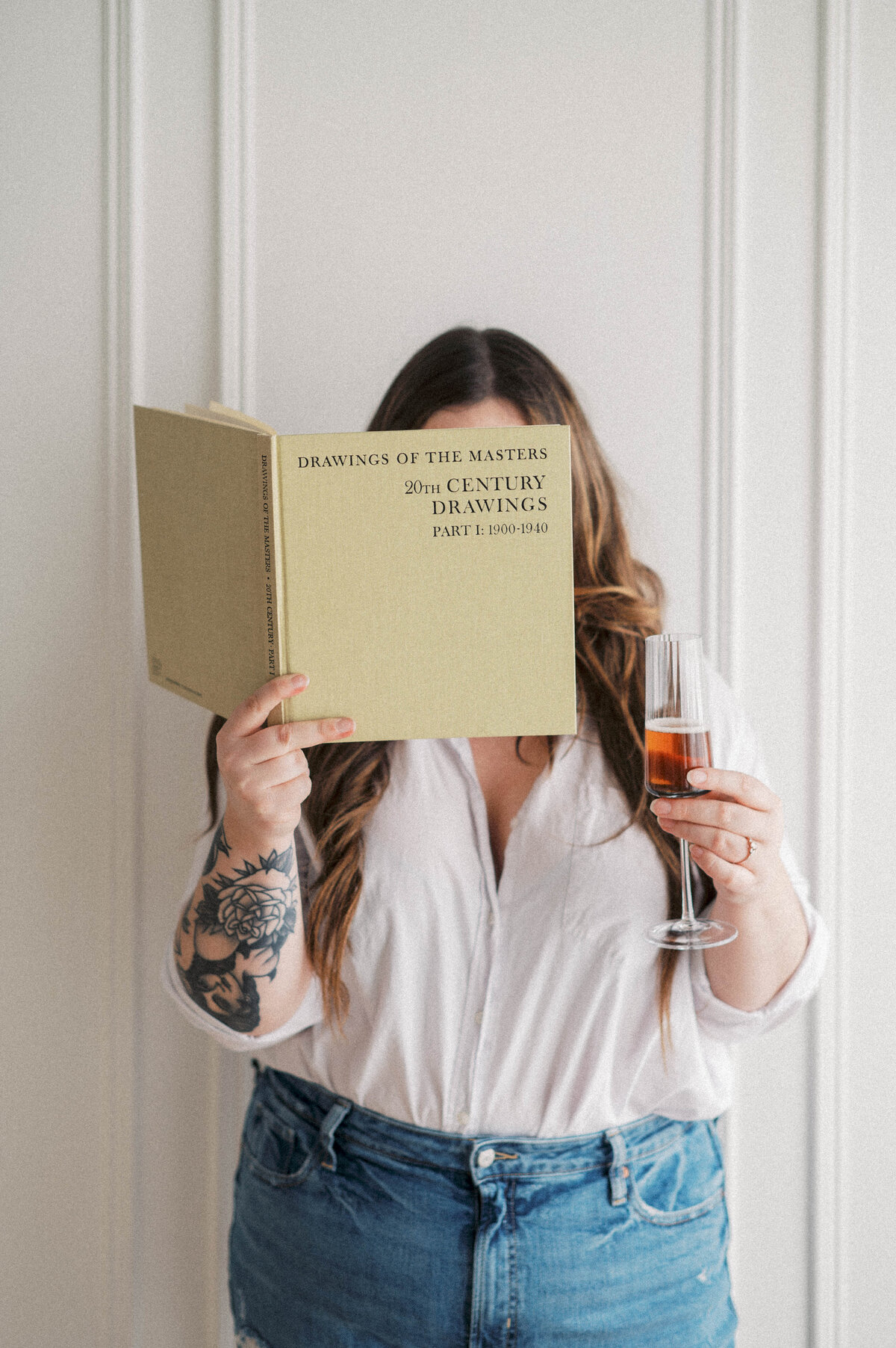 Woman holding art book infront of her face with glass of wine in the other hand