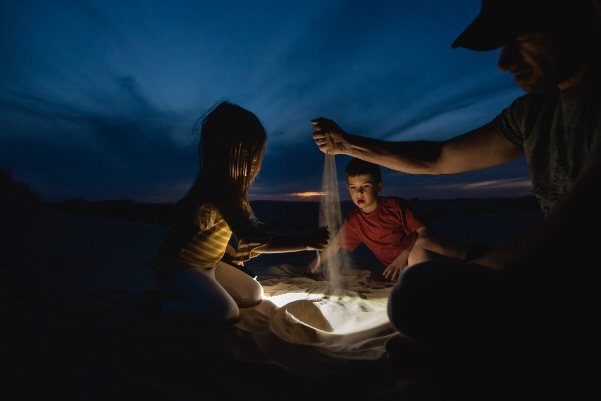 A blue hour night portrait of a family playing in the sand, lit only by a flashlight.