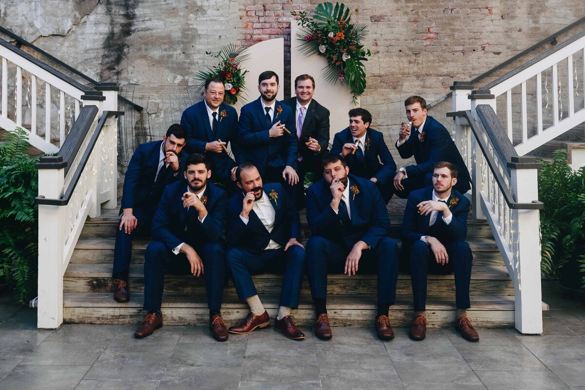 A group of ten men in blue suits pose playfully on wooden stairs, some crouching and some sitting. Behind them is a brick wall with a green and orange floral arrangement.