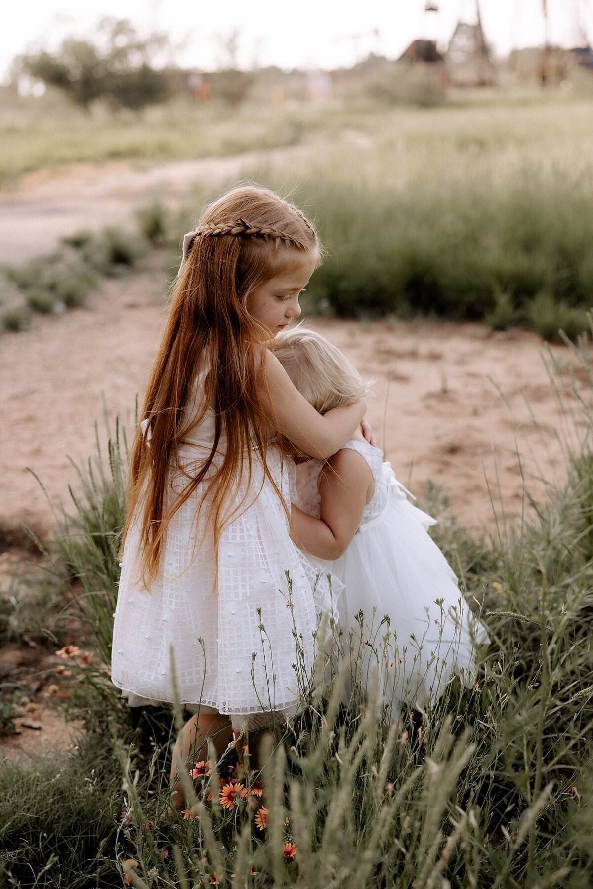two little girls standing in a field, older sister has her hands wrapped around the younger one giving her a hug.