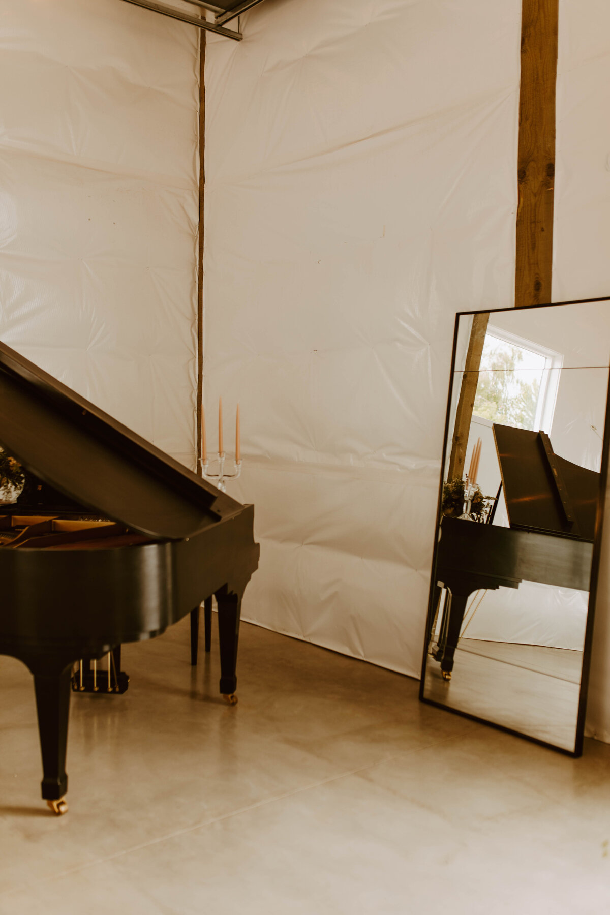 Vertical photo where a grand piano is half in frame and a large mirror rests against a wall to the right, reflecting it.