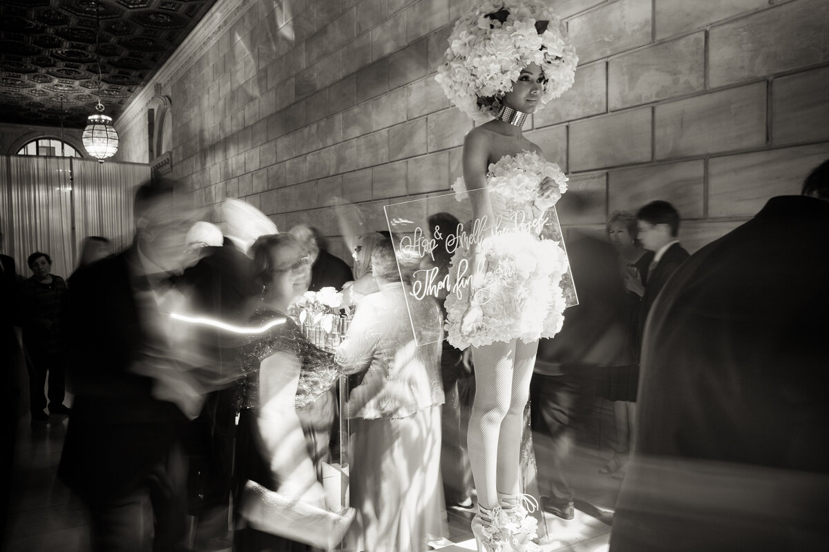 The captivating monochrome image was taken at The New York Public Library in New York City by the talented photographer Christian Oth. The photo captures a performer adorned in an extravagant costume, complete with large flowers, carrying a sign that reads Take a gift and thank you. Positioned on a pedestal, she exudes an air of mystery and elegance as passersby in blurred motion traverse the historical building with its grand high ceilings and elaborate walls. The scene beautifully merges artistry with the bustling energy of a busy day in the city.  Additionally, East West Films can be credited as Videographers for this information.