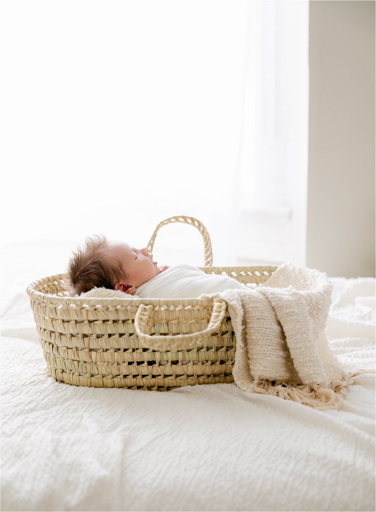 Newborn baby with brown hair is swaddled in white, laying in a moses basket on a bed in front of a large window during an Indianapolis newborn photography session.