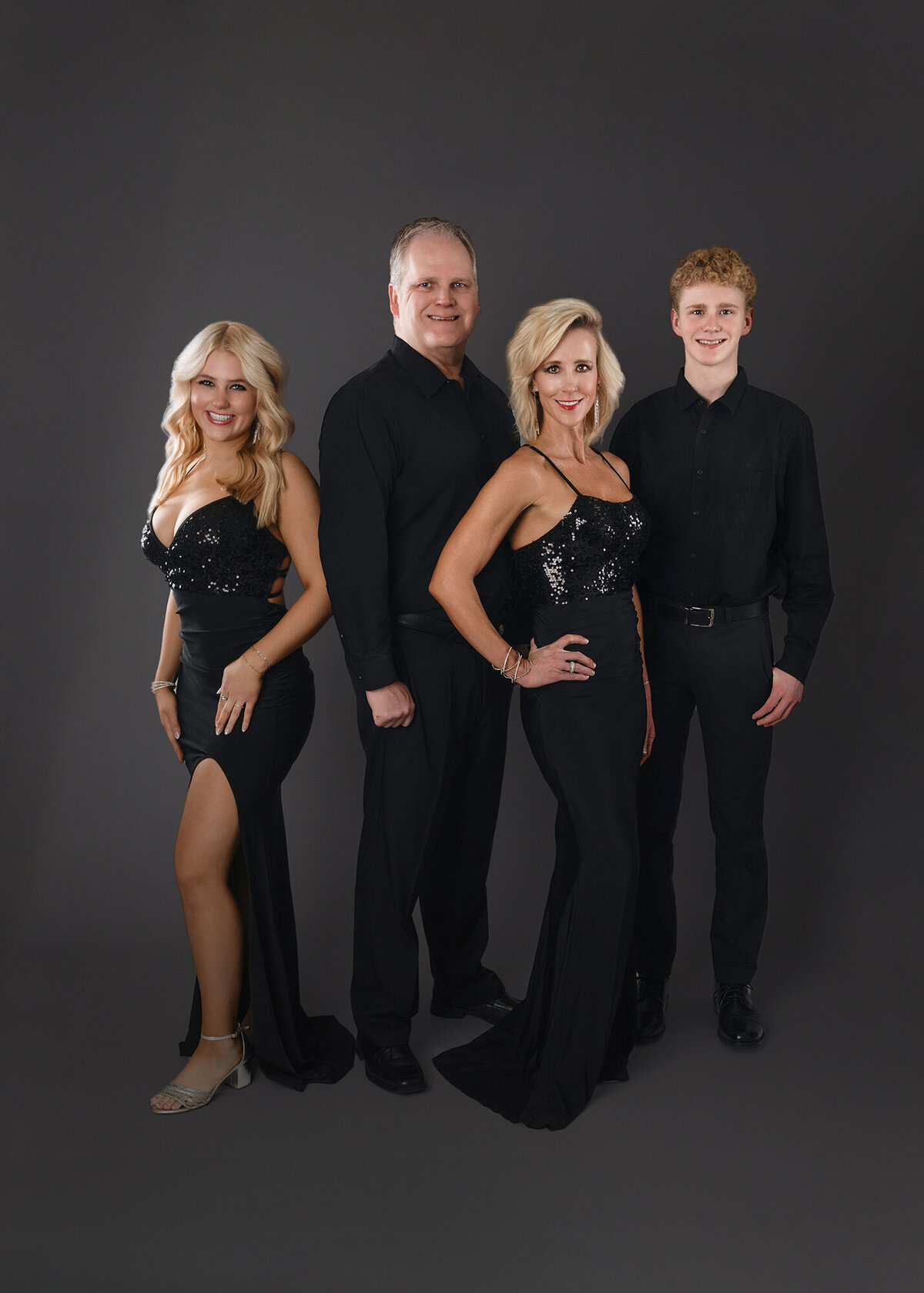A family of four, including two adults and two younger individuals, is dressed in formal black attire and standing against a dark grey background.