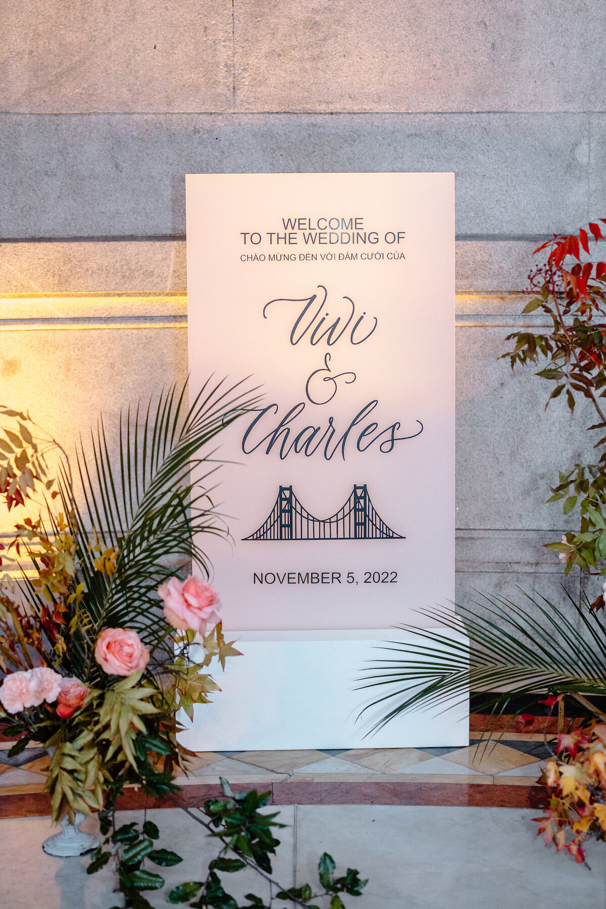 Black calligraphy lettering on a white frosted acrylic welcome sign, surrounded by florals, with a drawing of the golden gate bridge