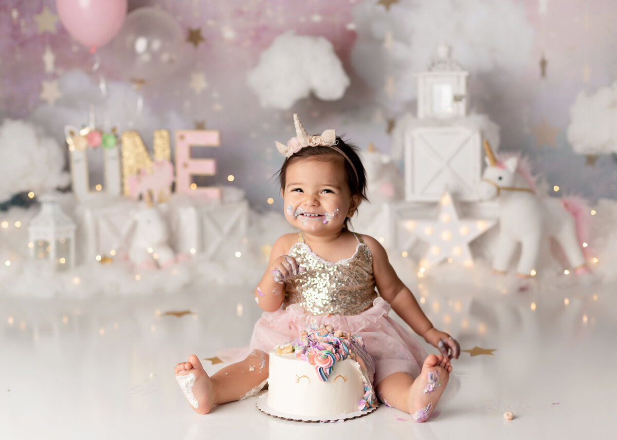 Unicorn themed cake smash with top West Palm Beach cake smash photographer. Baby girl is smiling at the camera with a unicorn cake between her legs and has icing on her hands, feet, and face. She is wearing a glittery gold and pink tulle dress with a unicorn headband. In the background, there are unicorns, starts, clouds, and balloons.