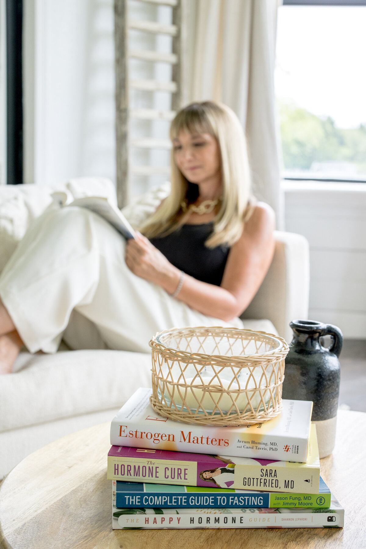 Brand photo of women in the background sitting on a white sofa in the living room reading a book and in front of her is a pile of books on a coffee table for her brand photo shoot