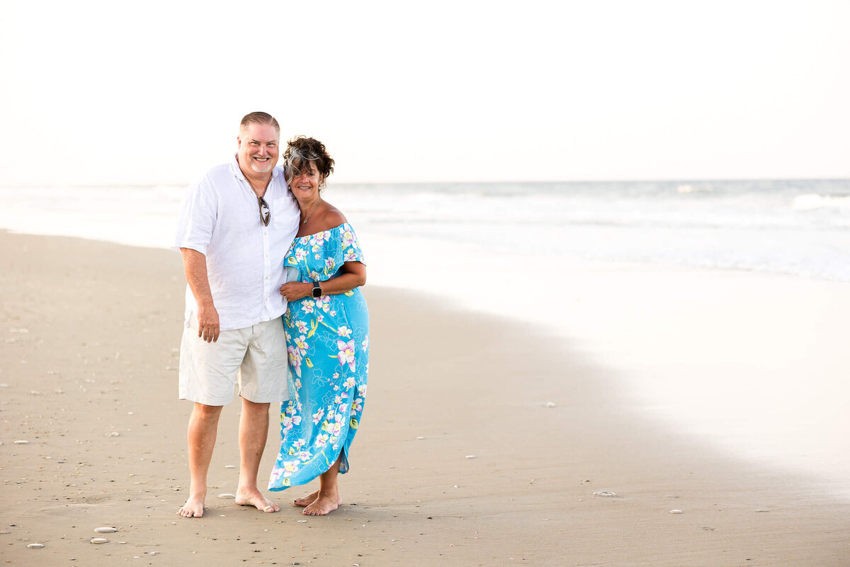 A man and a woman stand barefoot on a sandy beach, captured by a Fayetteville NC family photographer. The woman is wearing a blue floral dress, and the man is in a white shirt and beige shorts. They are smiling, with the ocean waves in the background.