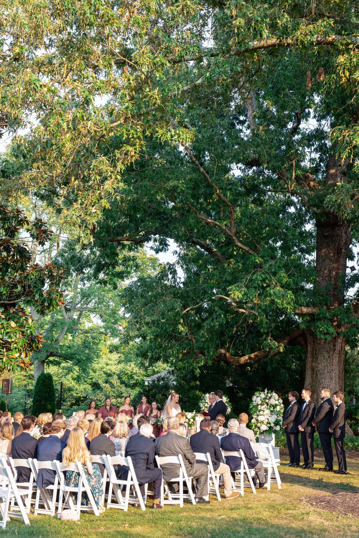 A wedding ceremony outdoors with guests seated on white chairs. The bridal party stands under a large tree as sunlight filters through the leaves. The setting is lush and green, creating a serene and intimate atmosphere.