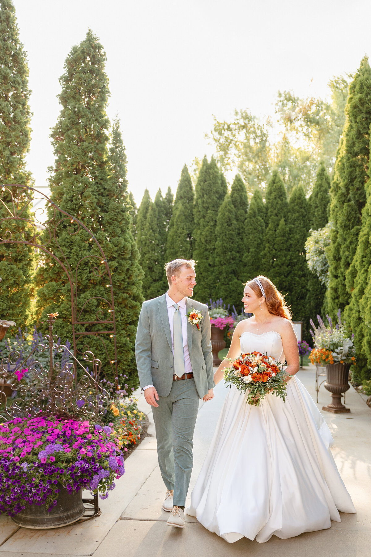 The newlyweds walk hand in hand down a garden pathway, sharing a joyful moment, with the bride’s flowing white dress and vibrant bouquet adding a touch of elegance.