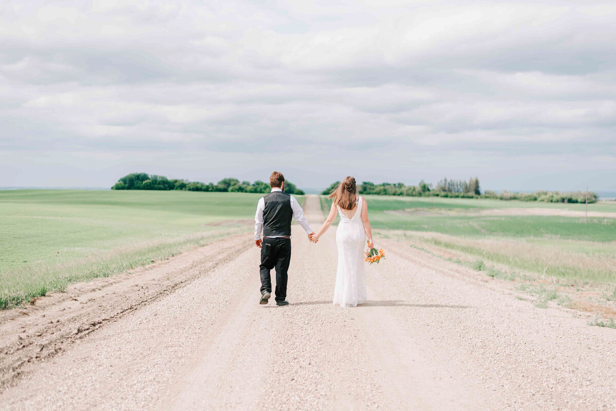 Bride and groom holding hands walking away from the camera on a gravel road by Allen, Saskatchewan.