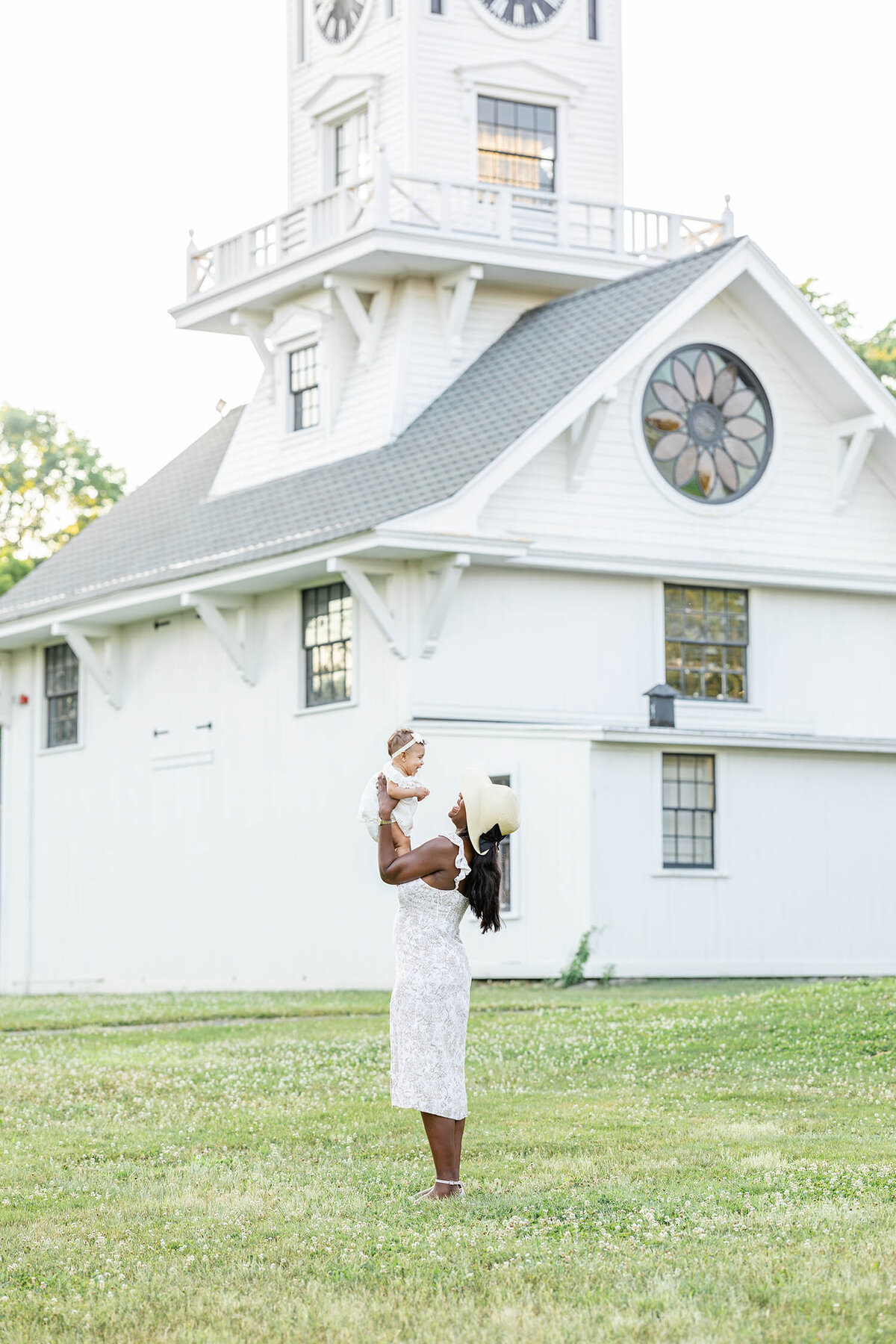 Woman wearing a dress and straw hat lifts baby in air during a mommy and me session