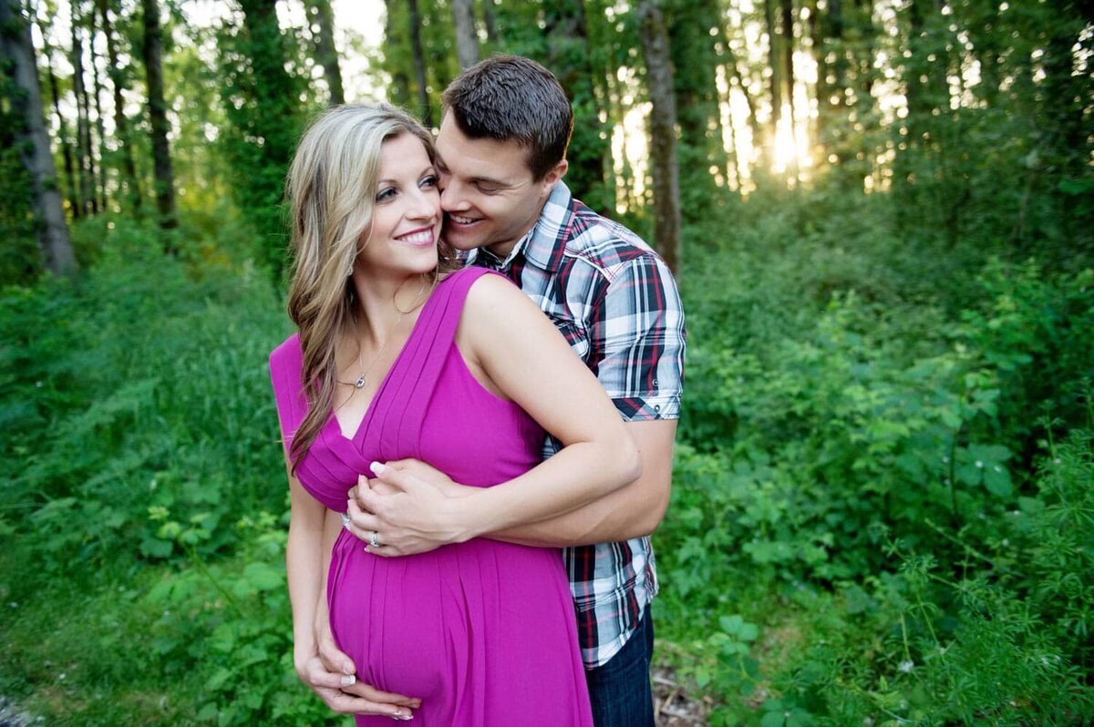 Expecting mother in a vibrant fuchsia dress with father-to-be hugging her from behind and holding hands in the forest.