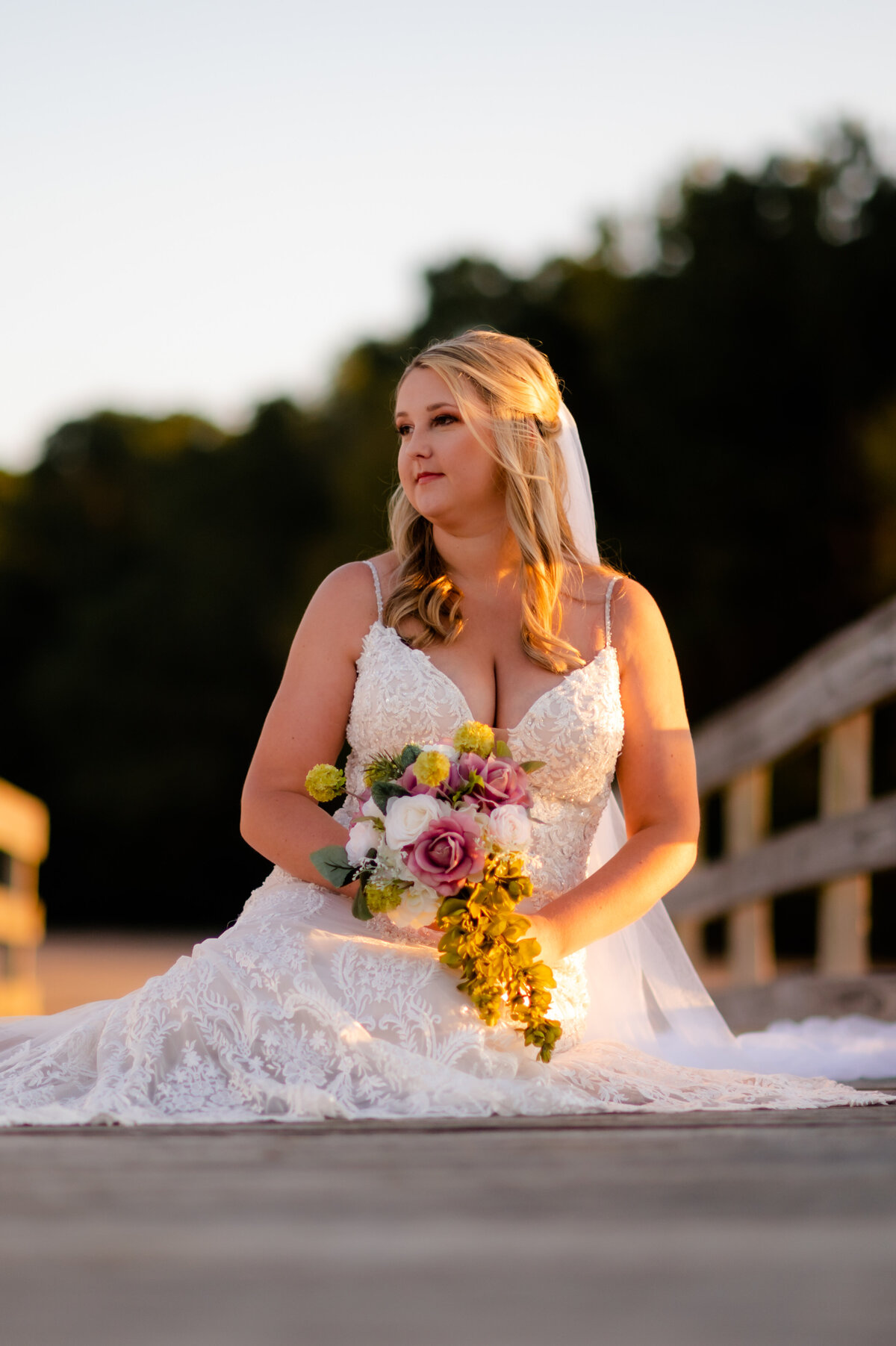 Best Little Rock wedding photographer photographs bride sitting on a wooden dock holding her bouquet while looking away from the sunset