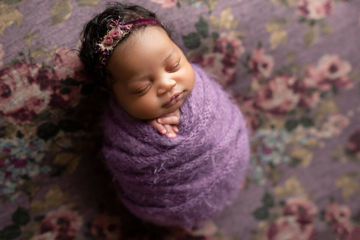 Black baby girl newborn photoshoot. Baby is wrapped in a purple swaddle with her fingers peeking out and is wearing a delicate floral headband. Baby is atop of a purple and dusty-rose floral stretch fabric.