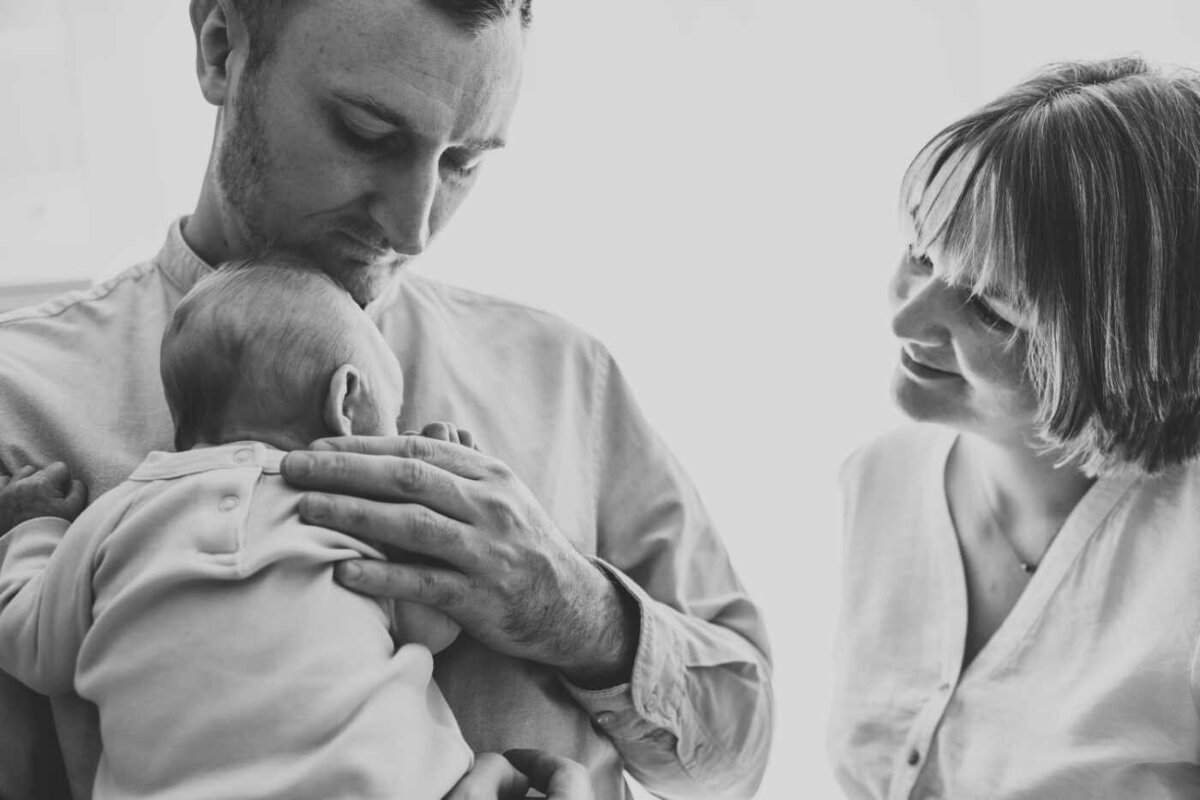 Ruua Hamdan Photography (UK): A black and white image of a father holding his baby, while the mother looks on with a smile.